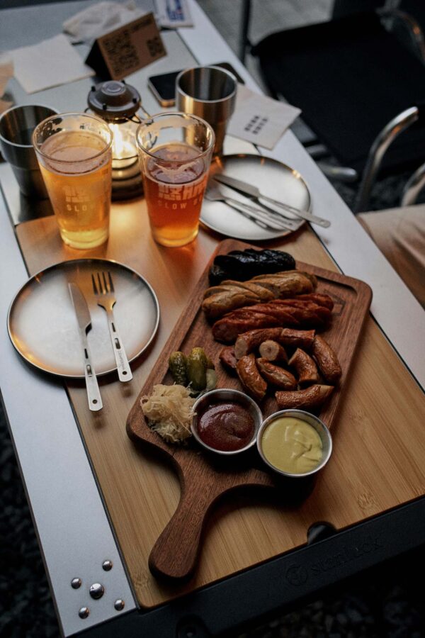 Charcuterie board with sausages, dips, and pickles next to glasses of beer.