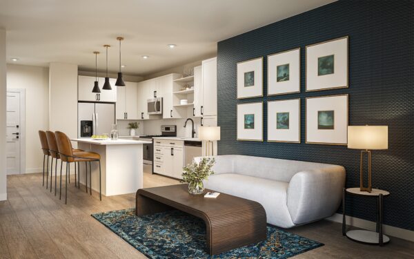 Interior rendering of a modern apartment showing a white kitchen with a breakfast bar and leather stools, adjacent to a living room with a gray sofa, dark textured wall with six framed art pieces, and a decorative blue rug.