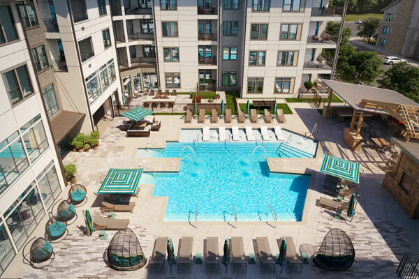 Aerial view of a turquoise swimming pool with four arched fountains. The surrounding deck includes gray lounge chairs, teal-striped umbrellas, woven egg chairs, and a covered outdoor kitchen area next to a modern multi-story apartment building.